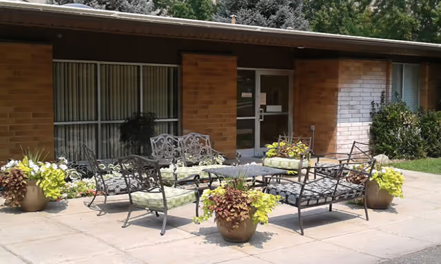 Outdoor patio area with metal chairs and benches with green cushions arranged around a small table. Several large potted plants with green and purple foliage are placed around the seating area. The patio is in front of a brick building with large windows and a glass door.