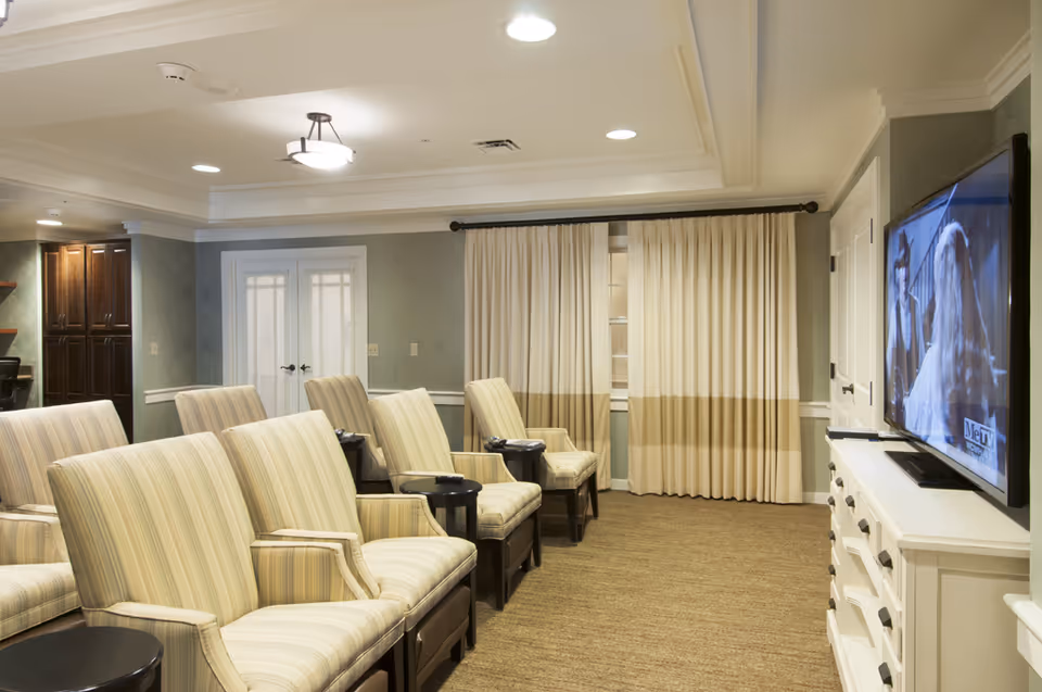 A cozy common area with several striped upholstered armchairs arranged in rows facing a wall-mounted flat screen TV. The room has beige carpeting, light-colored walls, and a set of closed curtains covering a window. There is a white cabinet below the TV and recessed ceiling lights providing illumination.