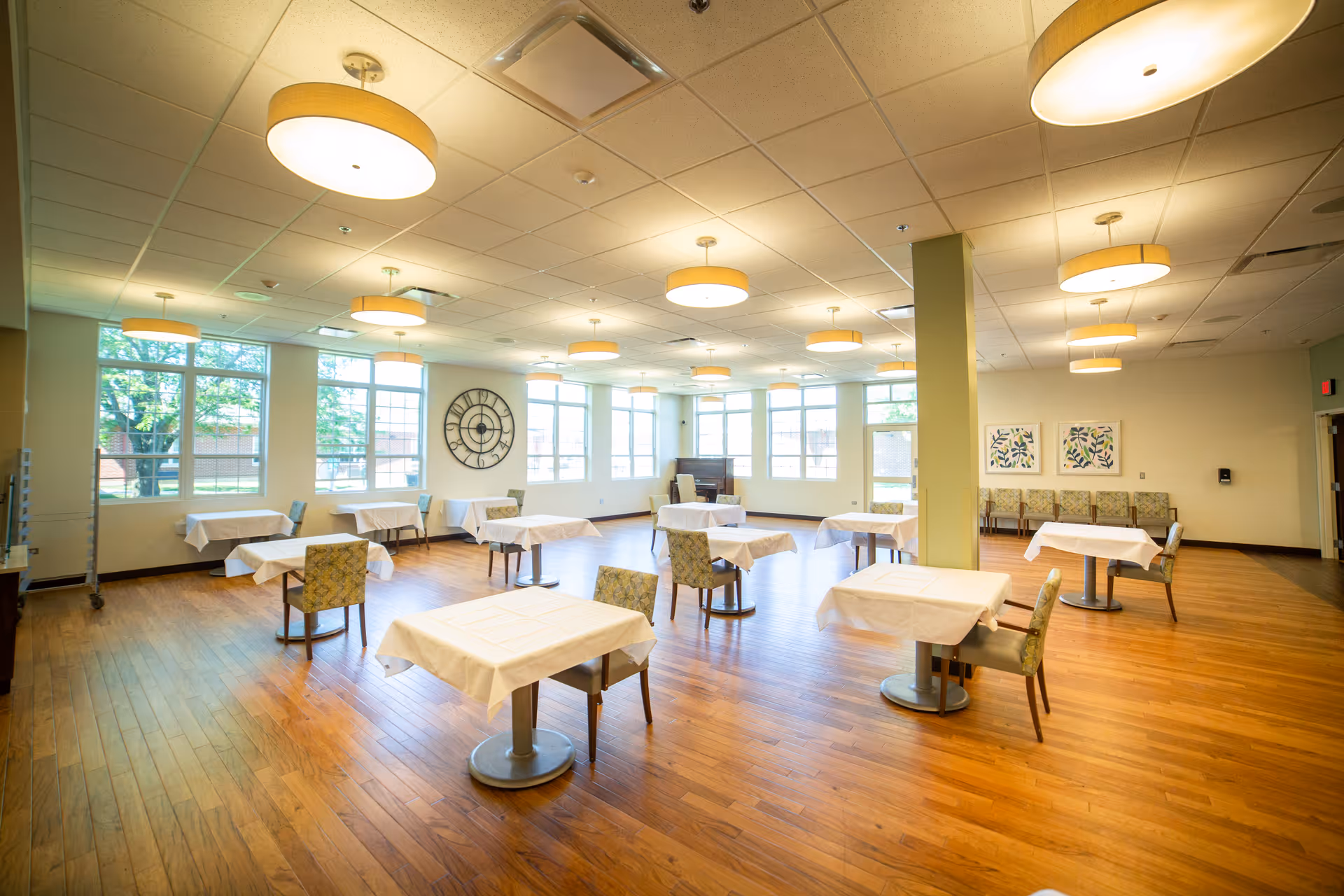 Spacious, well-lit dining room with multiple small tables covered in white tablecloths and chairs on hardwood floors.