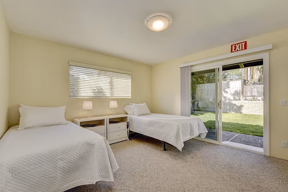 A bright bedroom with two single beds covered in white quilts, a small white nightstand with two lamps between the beds, a window with blinds on the left wall, and a sliding glass door leading to a grassy outdoor area with an exit sign above it.