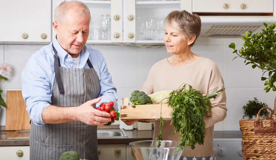 An elderly man and woman in a kitchen preparing fresh vegetables. The man is holding a bunch of radishes, and the woman is holding a wooden crate filled with various vegetables including broccoli and leafy greens. The kitchen has white cabinets and a countertop with a sink and some plants.