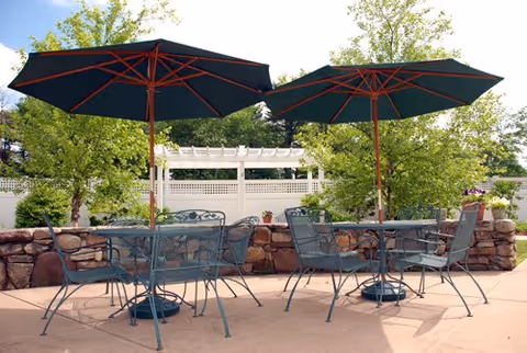 Outdoor patio area with two metal tables and chairs, each table shaded by a large green umbrella. The patio is surrounded by a low stone wall and greenery including trees and bushes.