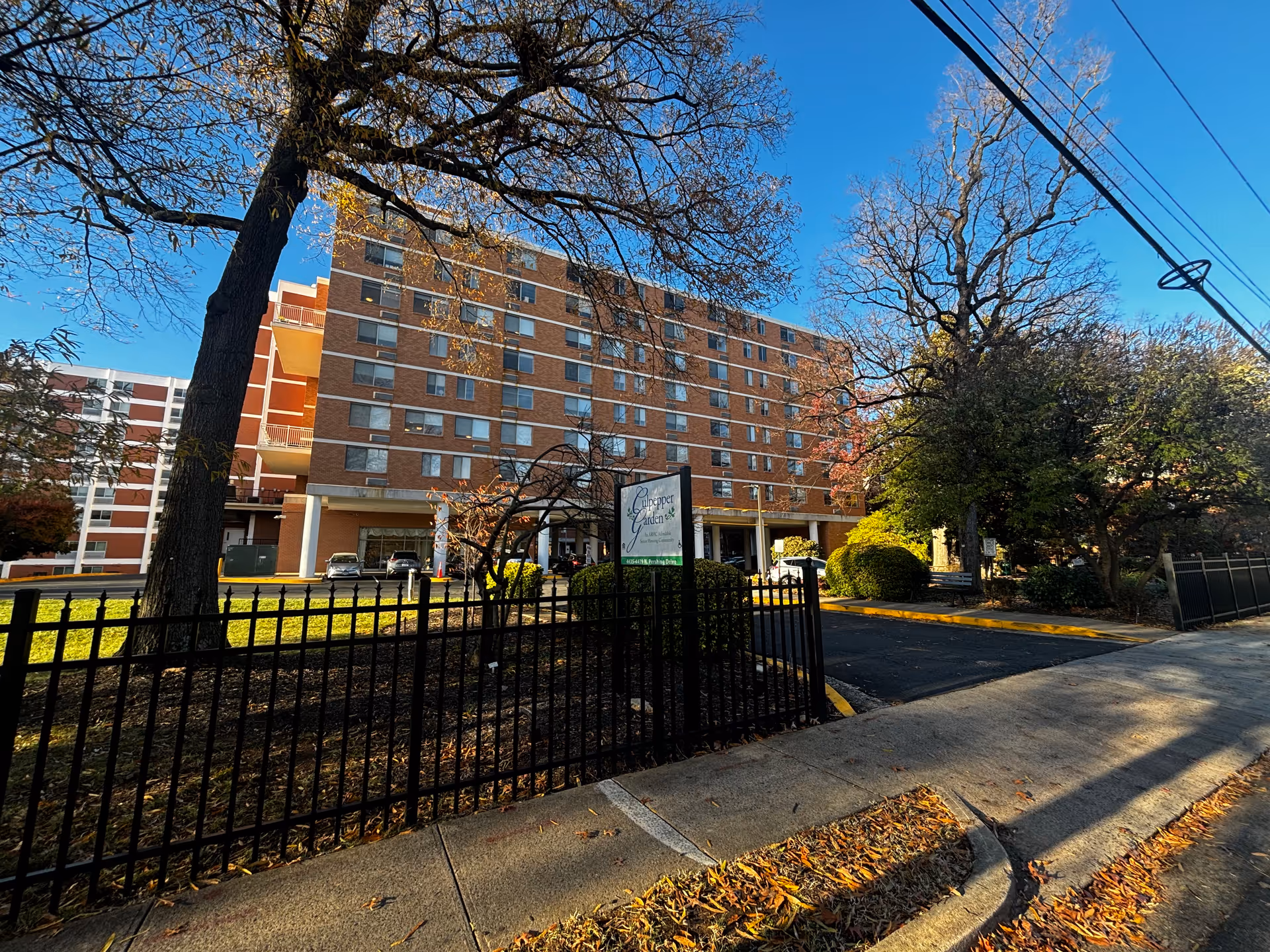 Exterior view of Culpepper Garden III, a multi-story brick building with several windows and balconies. The building is surrounded by trees with autumn leaves and a black metal fence. A sign with the facility name is visible near the entrance driveway. The sky is clear and blue.