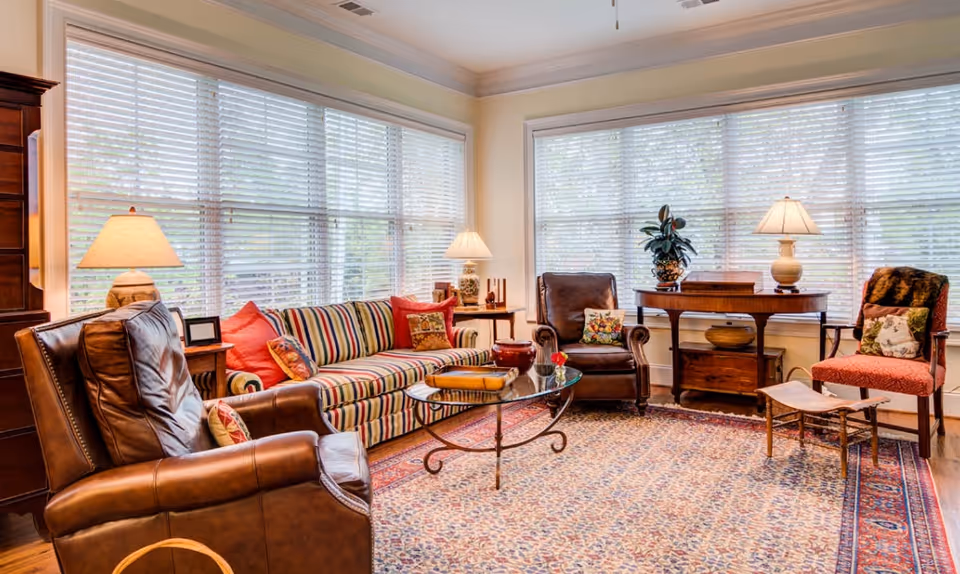 A cozy living room featuring a striped sofa with red cushions, two armchairs, a glass coffee table, and a wooden side table. Large windows with blinds allow natural light to fill the space, complemented by a lamp and decorative plants.