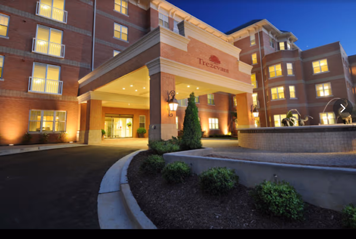 Exterior view of the Trezevant senior living facility at dusk, showing a multi-story brick building with illuminated windows, a covered entrance with columns, outdoor lighting, and landscaped bushes in front.
