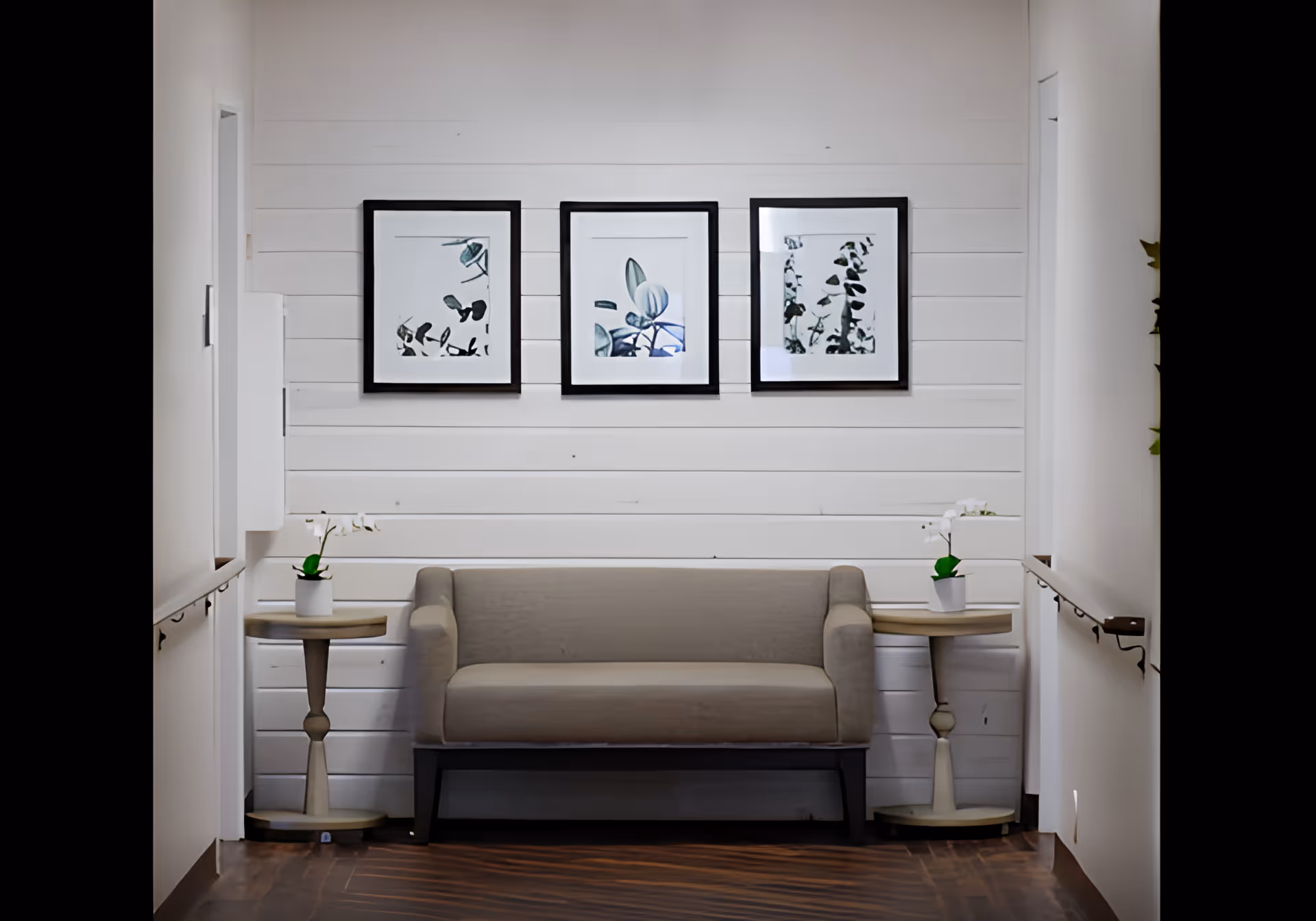 A small seating area in a hallway with a beige loveseat centered against a white shiplap wall. Above the loveseat are three framed botanical prints. On each side of the loveseat is a small round wooden side table with a white potted orchid. The floor is dark wood, and there are handrails on both sides of the hallway.