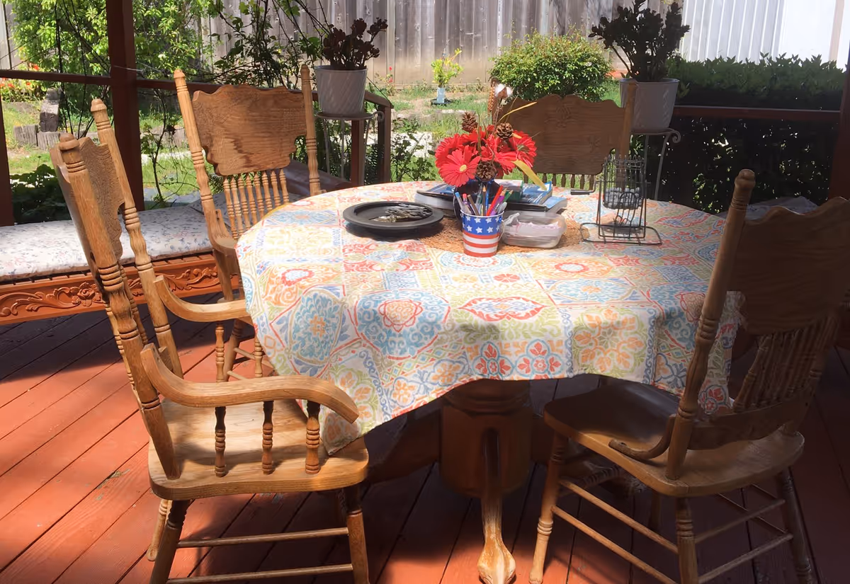 A round wooden table with a colorful patterned tablecloth on a wooden deck. The table is surrounded by four wooden chairs with carved backs and armrests. On the table, there is a small vase with red flowers, a container with pens and pencils, a tray with keys, and some other small items. In the background, there are potted plants and a wooden fence with greenery.