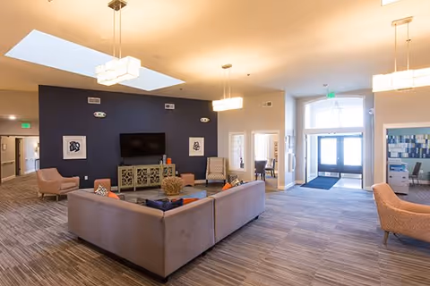 A spacious and well-lit living room area with a large beige sectional sofa, several armchairs, a flat-screen TV mounted on a dark blue accent wall, and modern light fixtures hanging from the ceiling. The room has large windows and a glass door letting in natural light, with a carpeted floor and a decorative console beneath the TV.