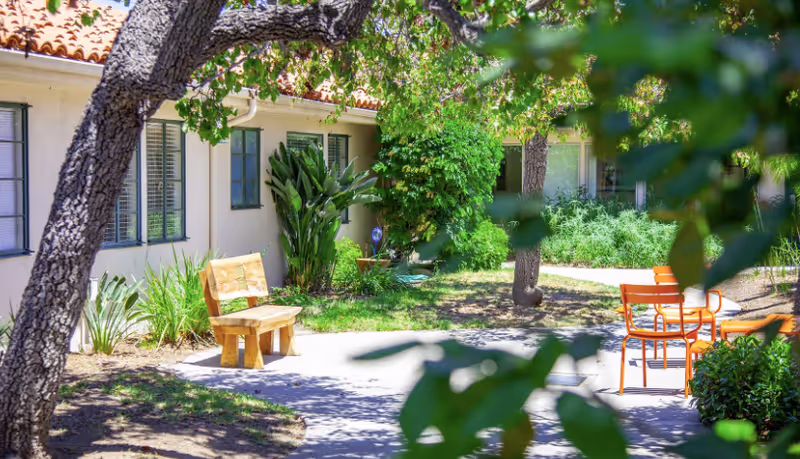 Sunny courtyard with a wooden bench, orange metal chairs and table, trees, and landscaping beside a single-story building.