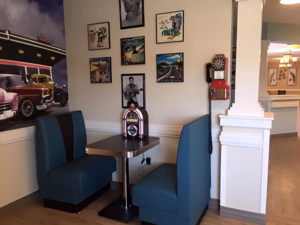 A retro-style dining booth with teal cushioned seats and a small table featuring a vintage jukebox. The wall behind the booth is decorated with framed pictures, including a black and white photo of Elvis Presley and other vintage-themed artwork. A red rotary wall phone is mounted on the adjacent wall. The setting has a nostalgic 1950s diner vibe.