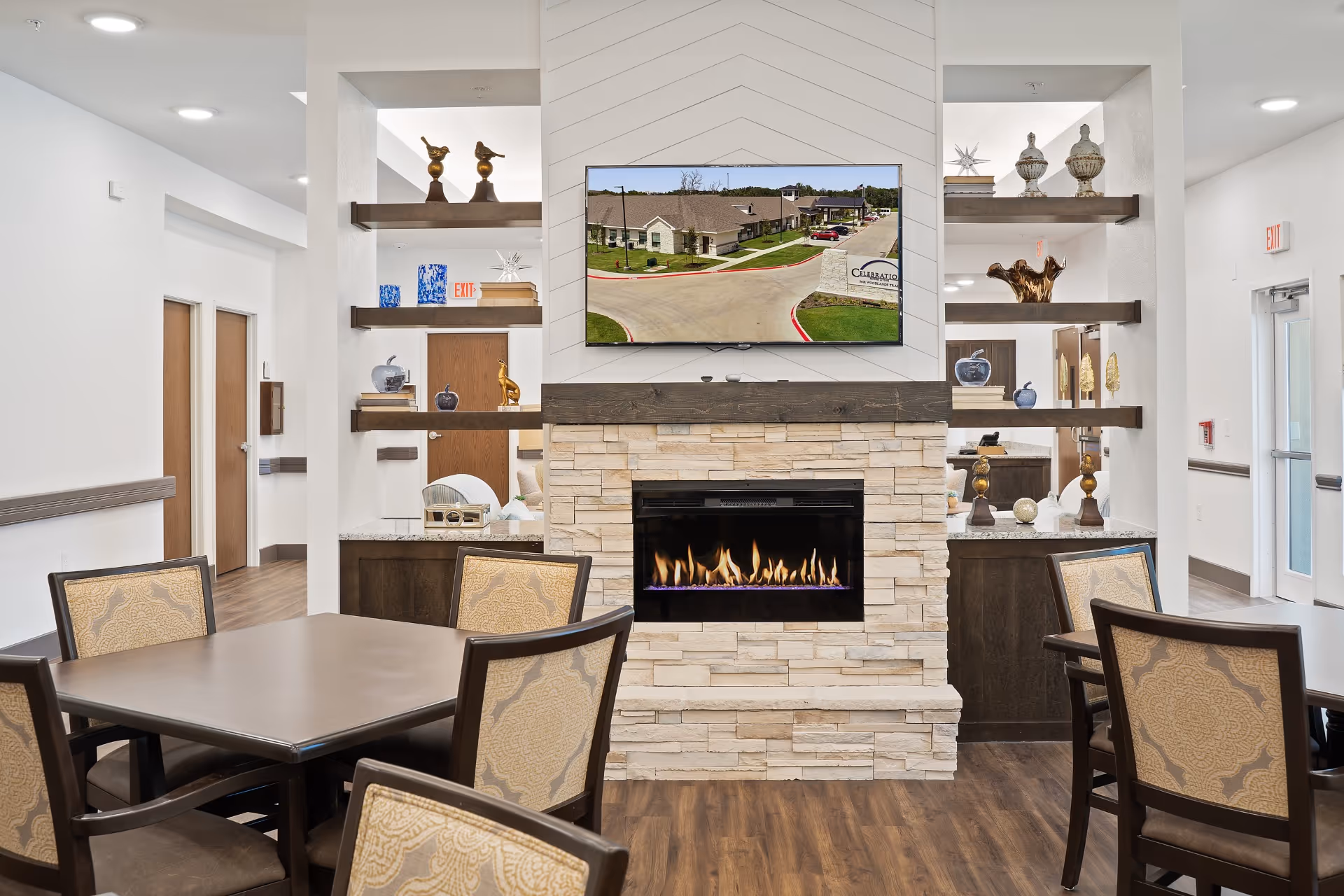 Interior view of a senior living facility common area featuring a modern stone fireplace with a flat-screen TV mounted above it displaying an aerial view of the facility. The room has wooden floors, several tables with upholstered chairs, decorative shelves with various ornaments, and white walls with wooden doors and exit signs.
