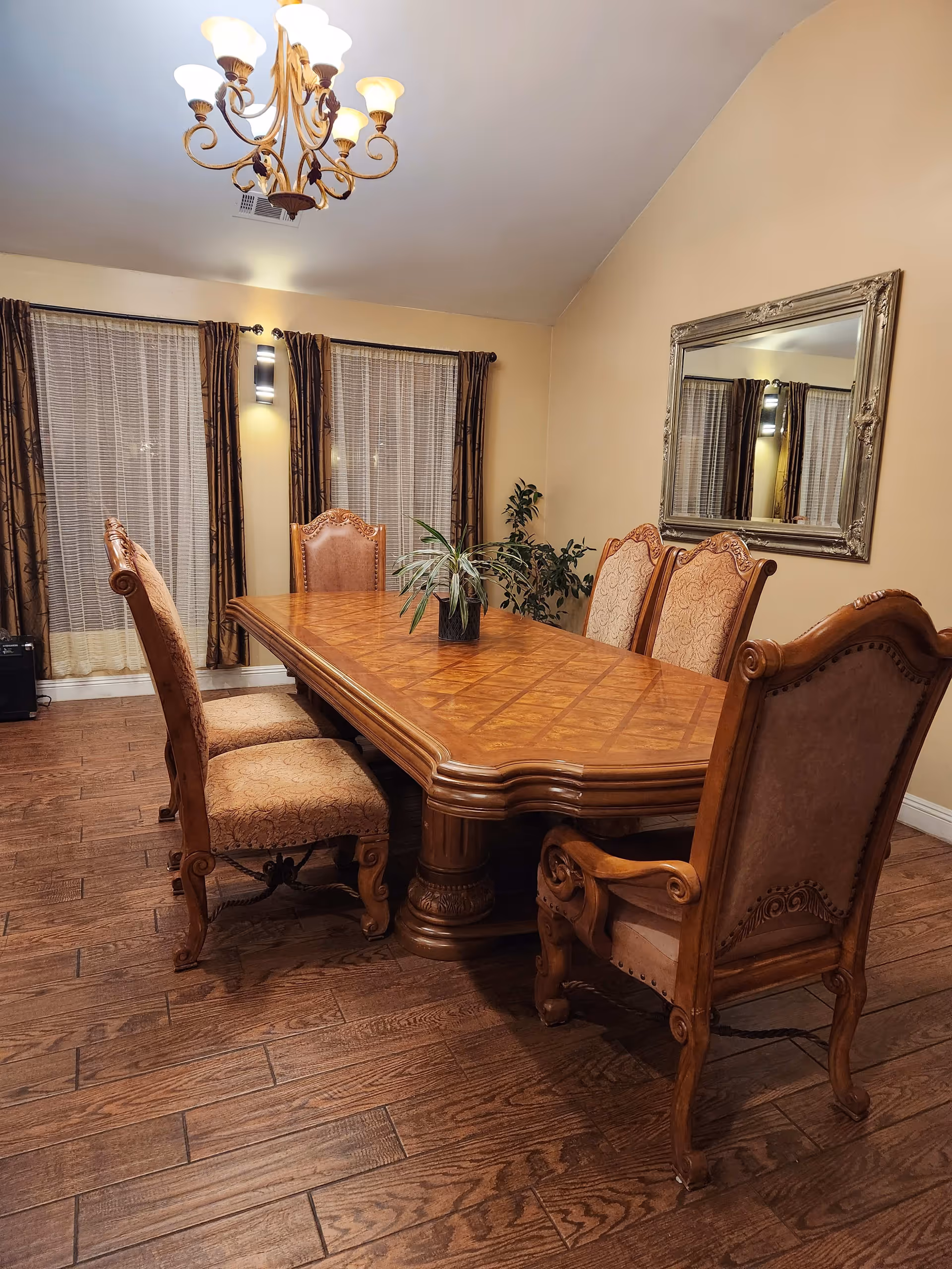 A dining room with a large wooden dining table surrounded by six upholstered chairs. The room has wooden flooring, two windows with curtains, a chandelier hanging from the ceiling, a large decorative mirror on the wall, and a potted plant in the corner.