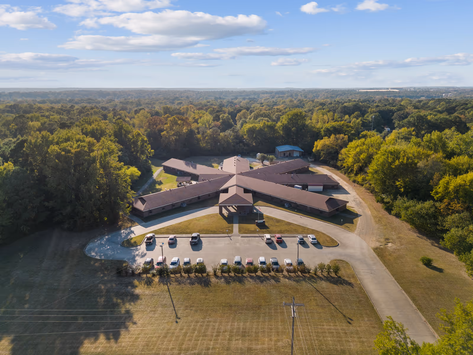 Aerial view of a star-shaped building surrounded by trees and greenery, with a parking lot in front containing several cars. The building is situated in a rural area with a clear sky and scattered clouds above.