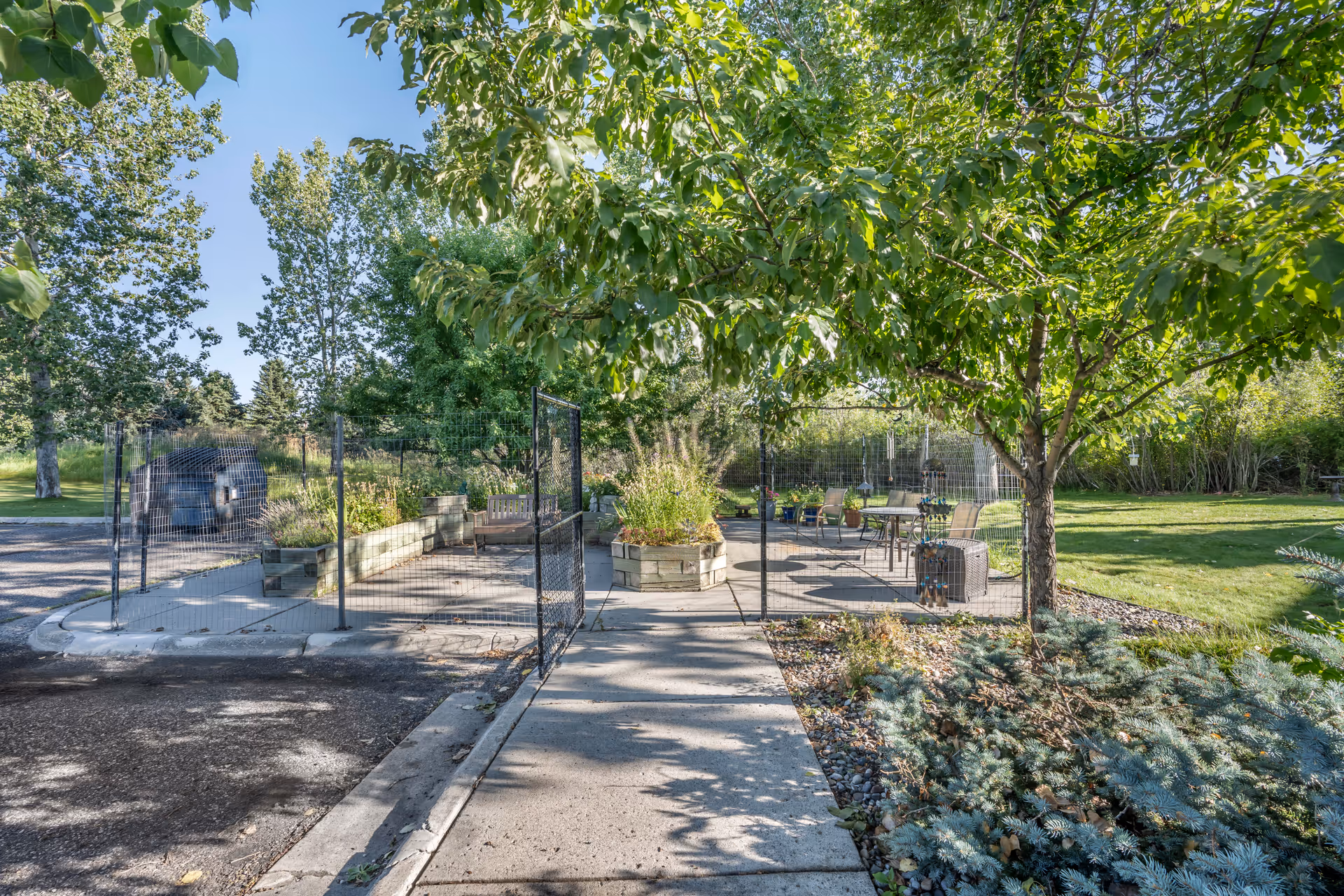 Outdoor patio area with a concrete walkway leading through a fenced garden space. There are trees providing shade, raised garden beds, and several chairs and tables arranged for seating. The surrounding area has green grass and various plants under a clear blue sky.