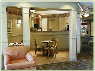 A cozy senior living common area with a small round dining table and chairs, an upholstered armchair, and a reception counter framed by columns.