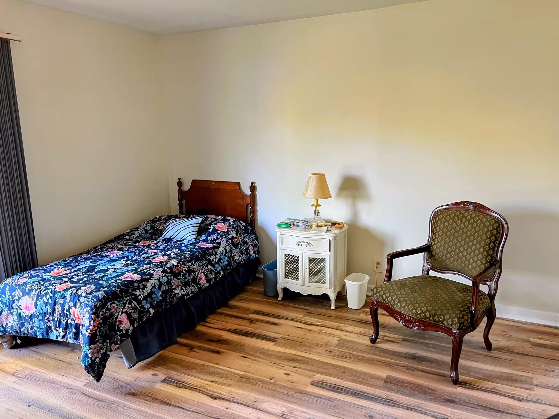 A simple bedroom with a single wooden bed covered with a floral patterned comforter and a striped pillow. Next to the bed is a small white nightstand with a lamp, some books, and a few items on top. There is a green upholstered wooden chair to the right of the nightstand. The room has wooden flooring and plain light-colored walls.
