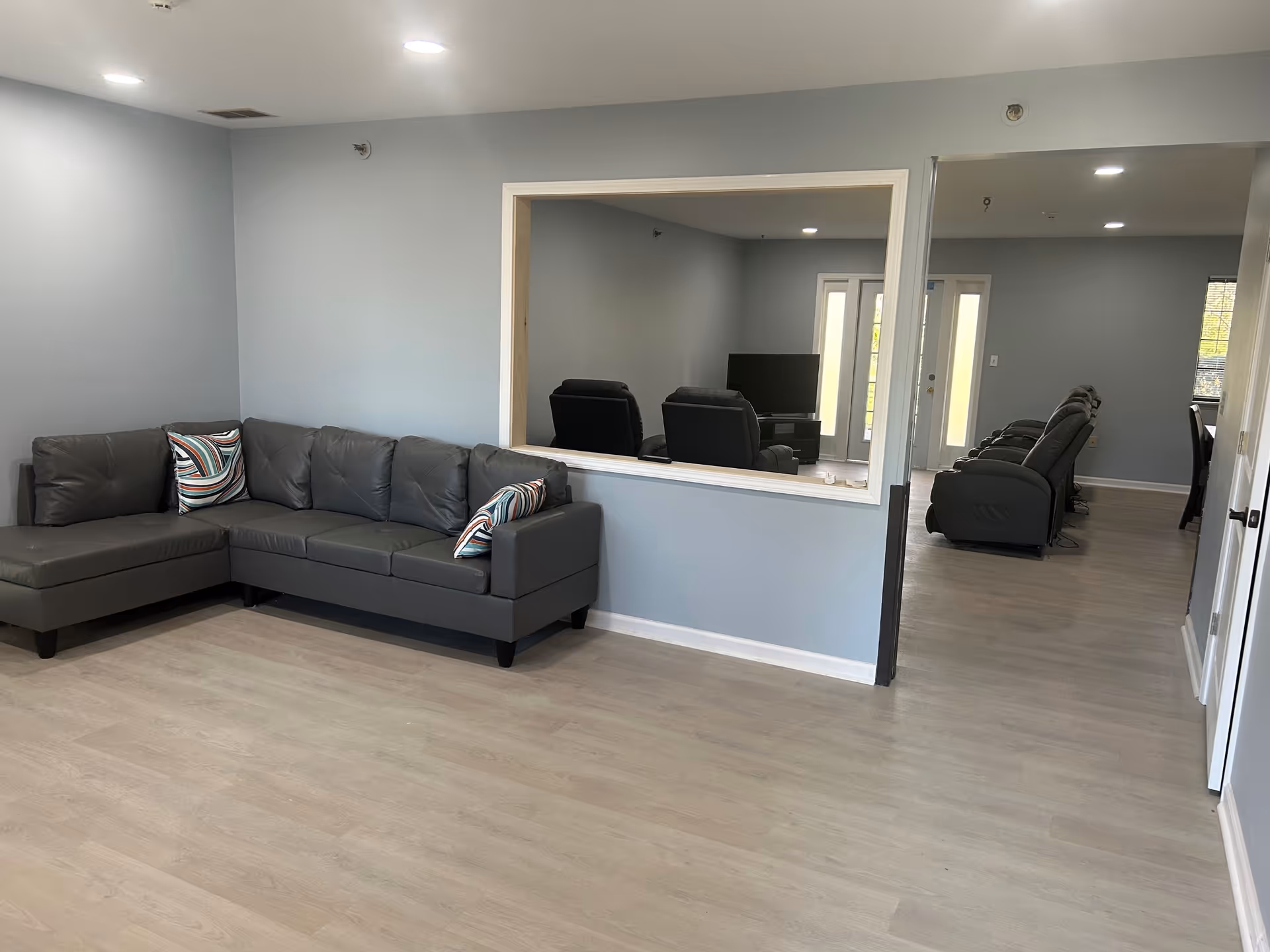 Spacious communal living room with a gray L-shaped couch, recliners and a TV visible through a pass-through window.