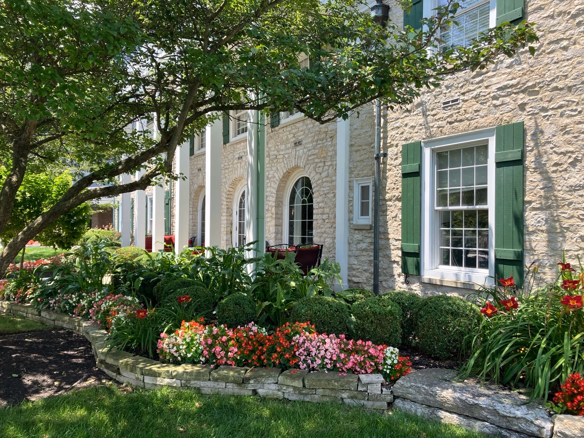 Stone building exterior with green window shutters and white columns, surrounded by a well-maintained garden featuring various green plants and colorful flowers under a tree providing shade.