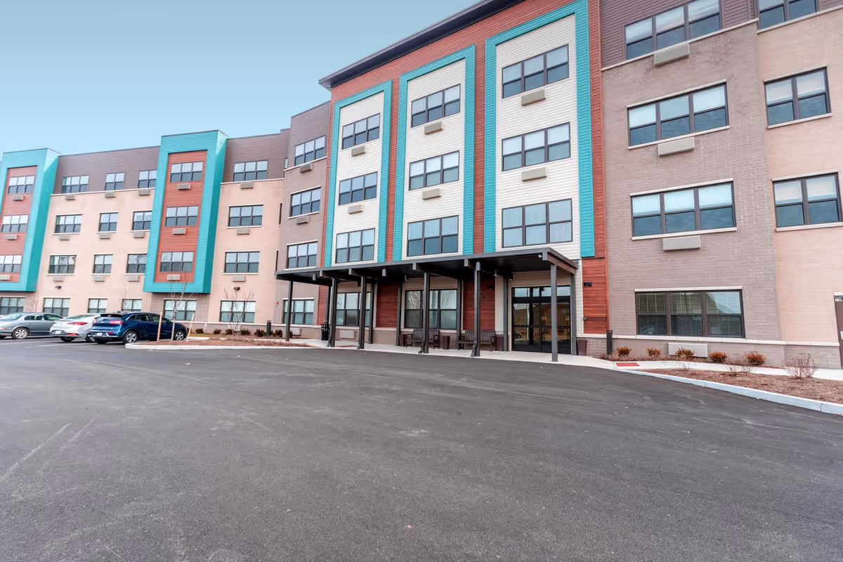 Exterior view of a multi-story senior living facility building with a covered entrance, several windows, and a parking area with cars in front.