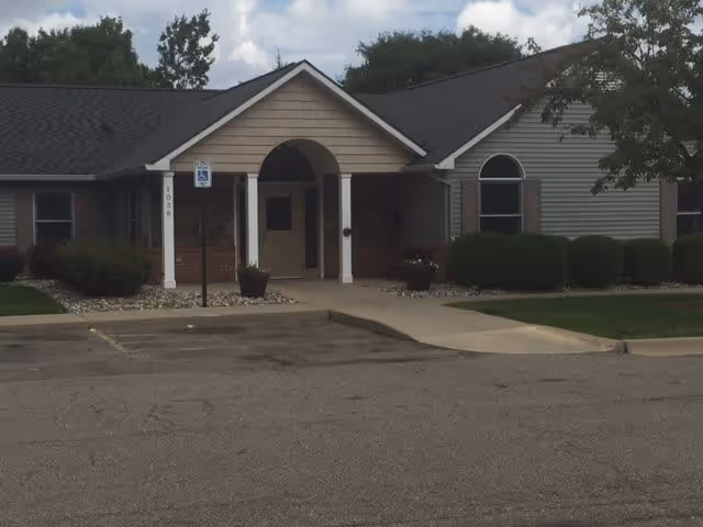 Front exterior view of a single-story assisted living facility building with a covered entrance, bushes, and a parking area including a handicapped parking sign.