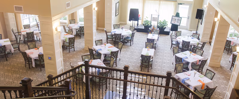 A spacious dining room with multiple tables covered in white tablecloths, each set with colorful napkins and chairs arranged neatly around them. The room has large windows with vertical blinds allowing natural light to fill the space. There are decorative columns and a carpeted floor, with a staircase railing visible in the foreground.