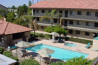 Outdoor swimming pool area with lounge chairs, tables with umbrellas, and a small building with a tiled roof. In the background, there is a three-story residential building with balconies and palm trees around the pool area.