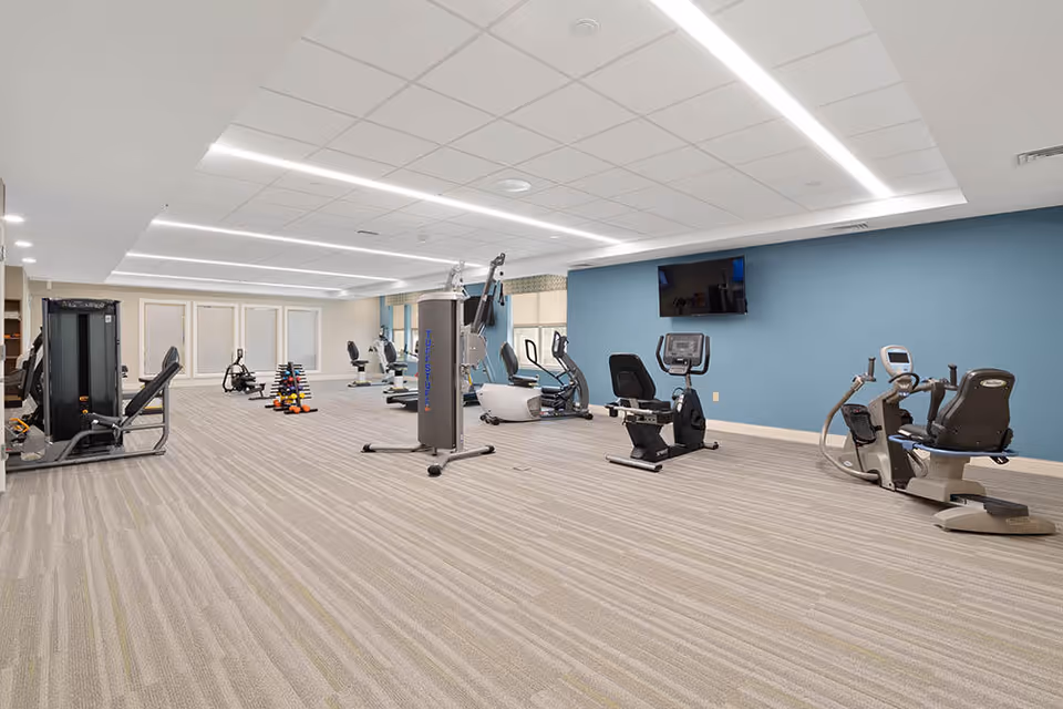 A spacious fitness room with various exercise equipment including stationary bikes, weight machines, and free weights. The room has a blue accent wall with a mounted flat-screen TV, beige carpet flooring, and bright overhead lighting.