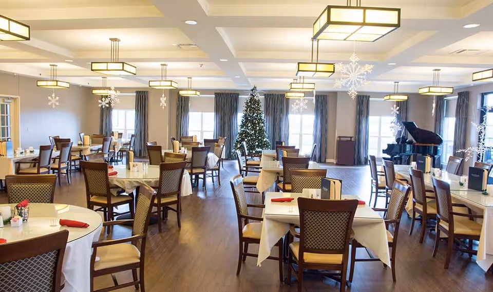 A spacious dining room decorated for the holidays with a Christmas tree and hanging snowflake ornaments. The room features multiple tables covered with white tablecloths, each surrounded by wooden chairs with cushioned seats. Large windows with curtains allow natural light to fill the room, and a grand piano is visible in the background.