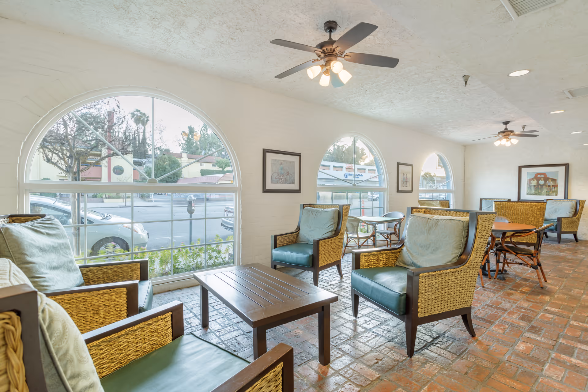 A bright and airy common area with large arched windows letting in natural light. The room features wicker armchairs with blue cushions arranged around wooden coffee tables, ceiling fans with lights, framed artwork on the walls, and a brick floor. Outside the windows, cars and trees are visible.
