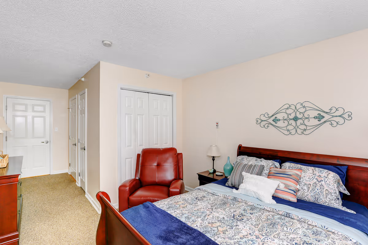 A bedroom in a senior living facility with a wooden bed frame, a bed made with patterned bedding and multiple pillows, a red leather armchair, a nightstand with a lamp and decorative items, and a closet with white double doors. The walls are light beige and there is a decorative metal wall art above the bed.
