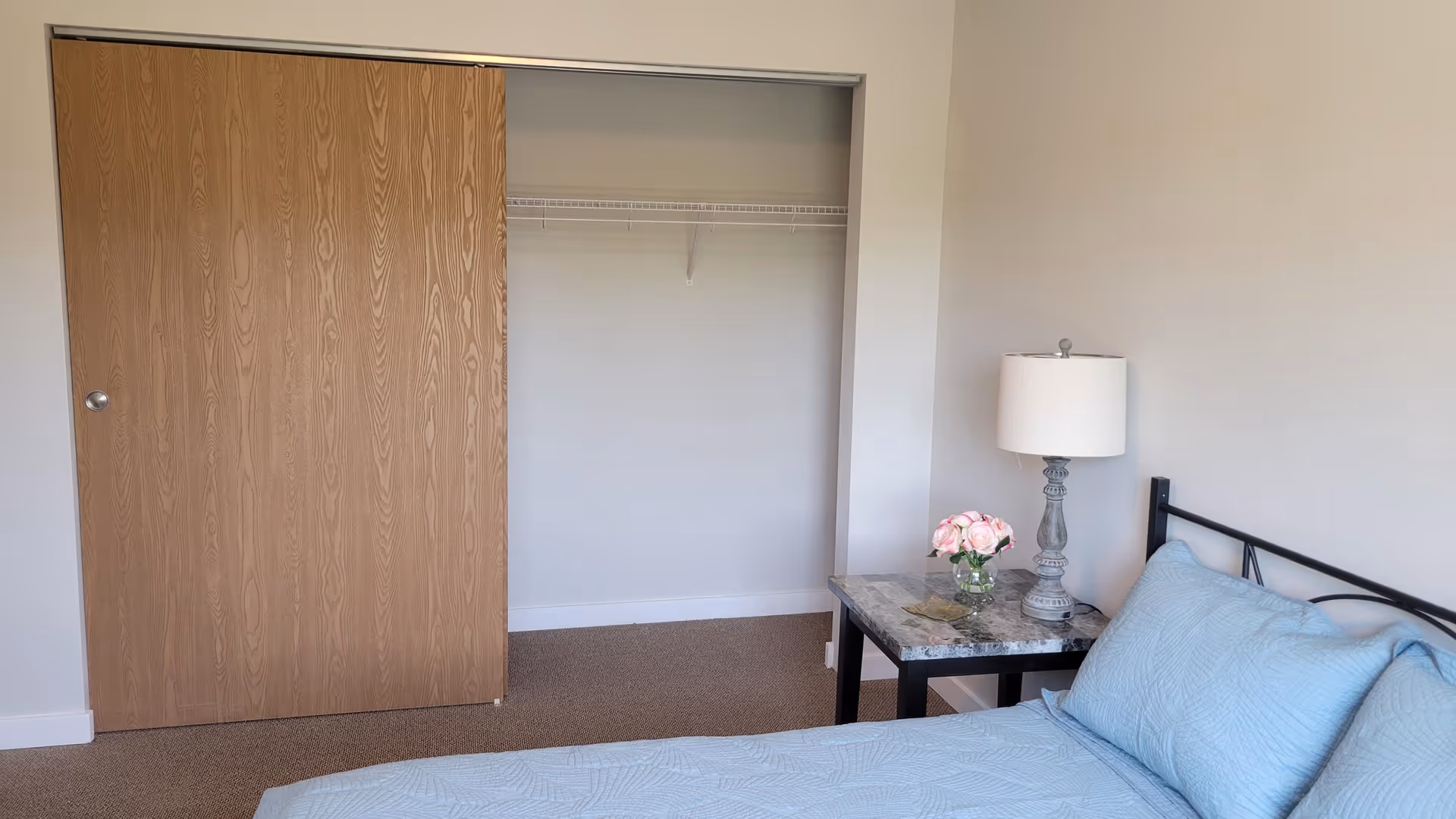 A bedroom with a bed covered in light blue bedding, a bedside table with a marble top holding a lamp and a small vase of pink flowers, and an open closet with a sliding wooden door and a wire shelf inside.