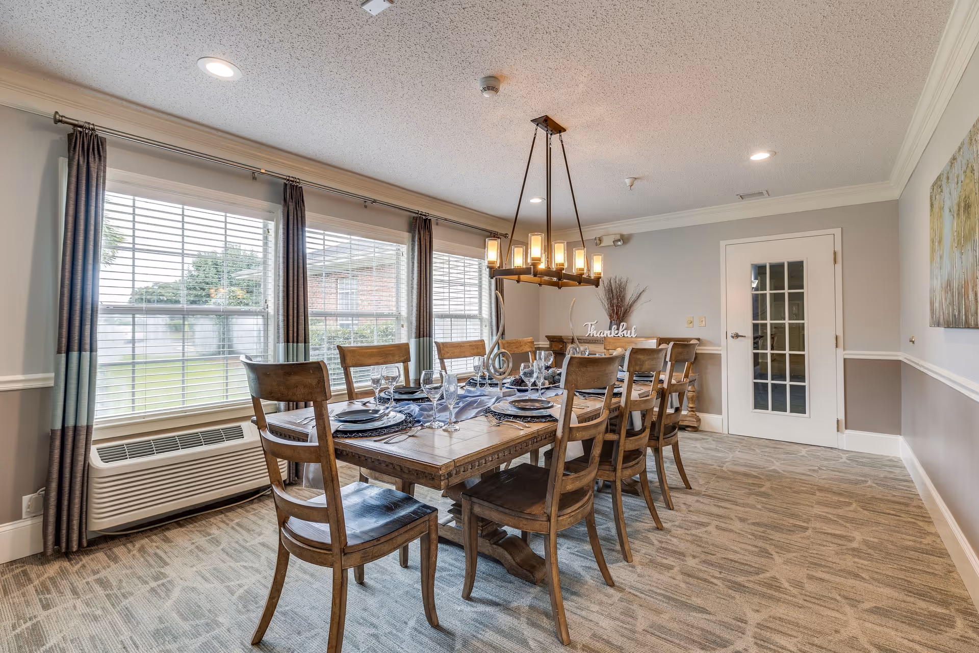 A dining room with a long wooden table set for eight people with plates, glasses, and silverware. The room has large windows with curtains, a modern chandelier hanging above the table, and a door with glass panels. The walls are painted light gray with white trim, and there is a decorative piece on the far wall.