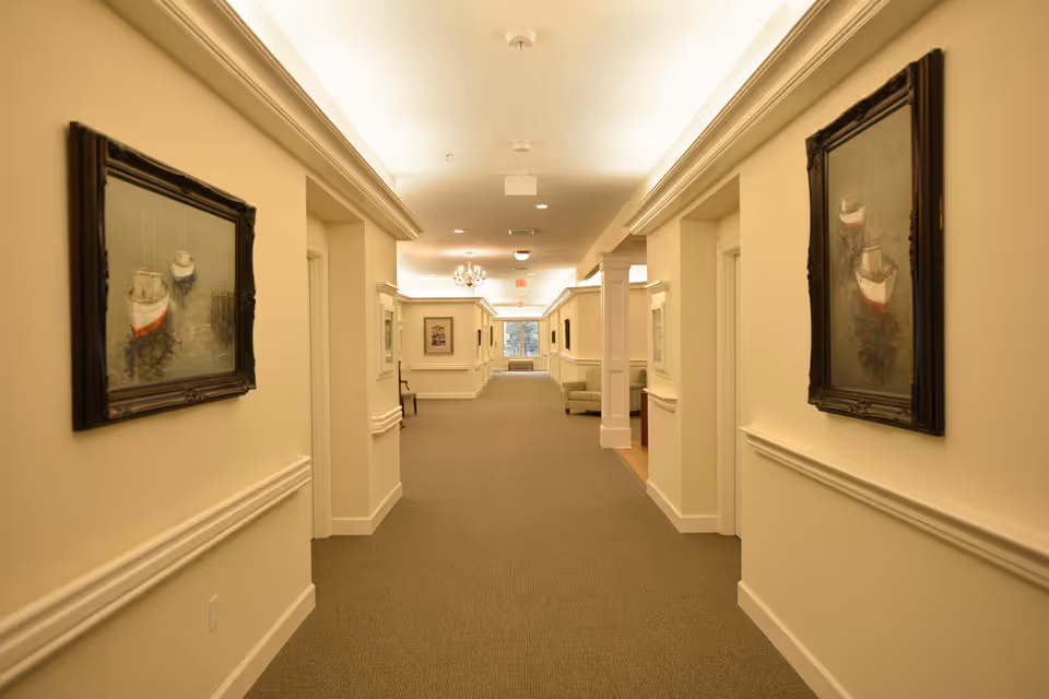 A well-lit hallway in a senior living facility with beige walls, carpeted floor, and framed paintings of boats on both sides. The hallway has white trim and crown molding, with a chandelier and recessed lighting on the ceiling. There are chairs and a small table visible further down the hall near a window.