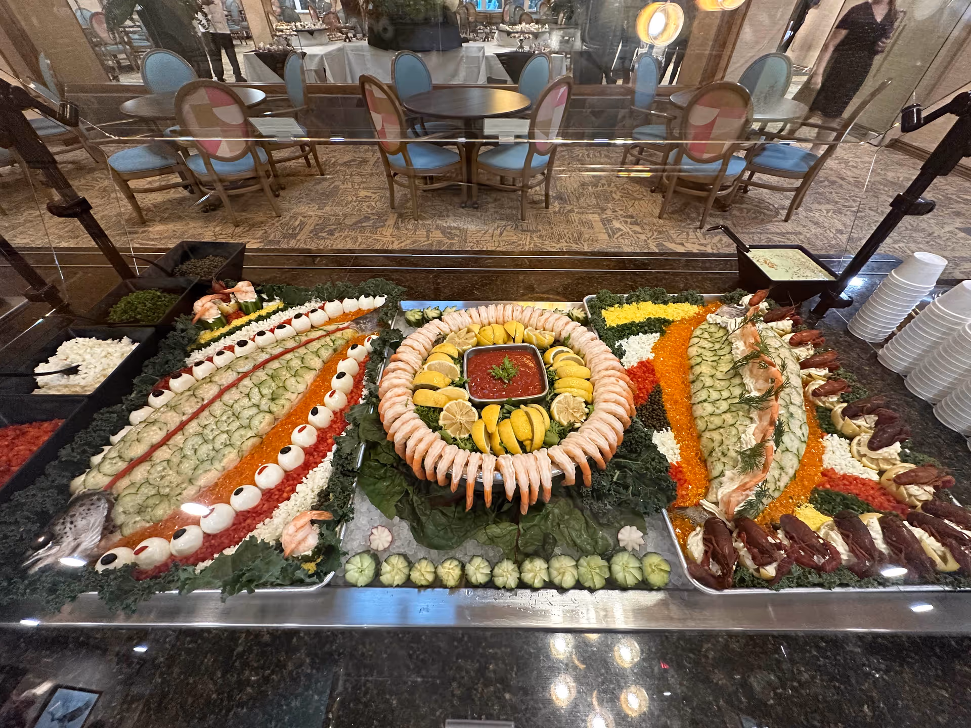 A large buffet seafood display featuring a ring of shrimp, garnishes, and assorted salads behind a sneeze guard in a dining room.
