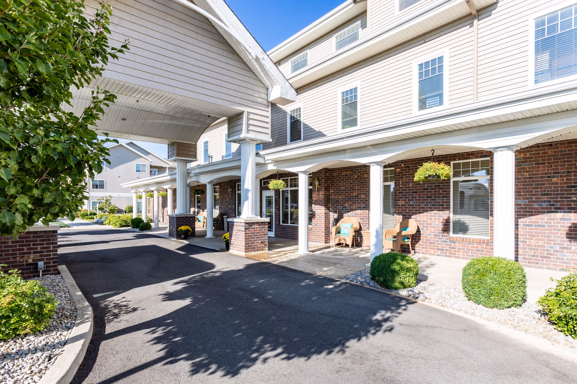 Exterior view of Broadway Court Estates showing a covered driveway entrance with white columns and brick walls. There are chairs with cushions on the porch area and hanging plants above. The building has multiple windows and is surrounded by greenery and shrubs.