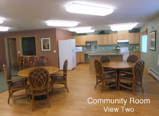 Community room with wooden round tables and chairs, a kitchen area with light wood cabinets, white refrigerator, stove, and microwave. The room has wood flooring and neutral-colored walls with framed pictures.
