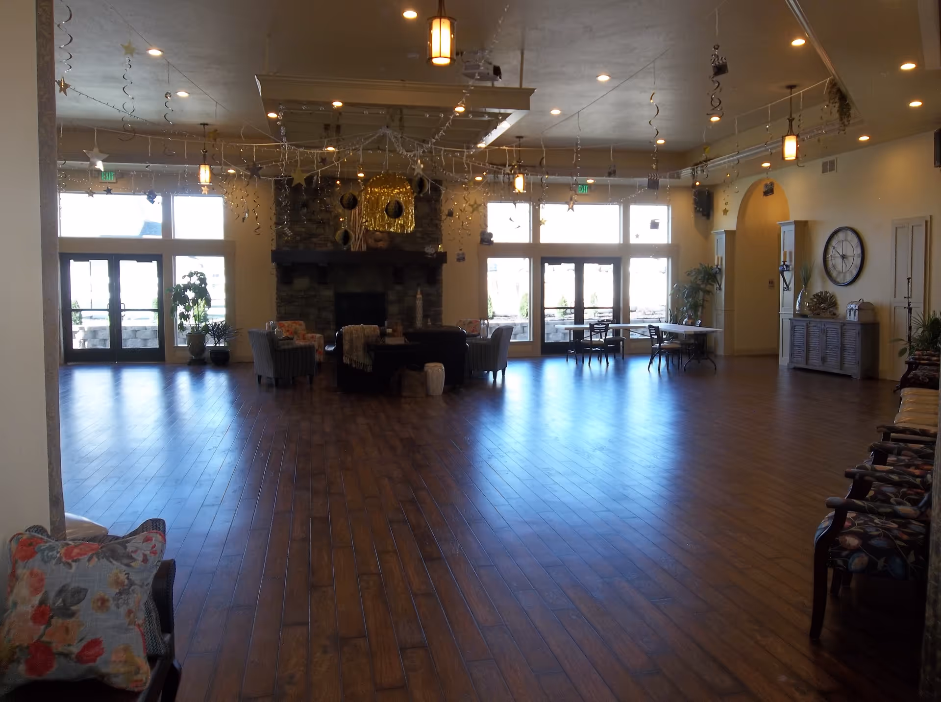 Spacious common lounge with wood floors, seating arranged around a central stone fireplace and large windows/doors at the far wall.