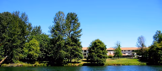 A low-rise residential building sits across a pond, partially hidden by trees under a clear blue sky.