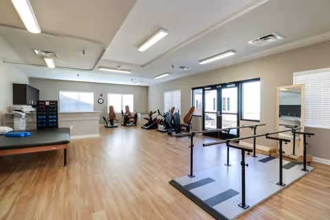 A spacious rehabilitation room with wooden flooring, exercise equipment including stationary bikes, parallel bars for walking practice, a treatment table, and large windows allowing natural light. The room has neutral-colored walls and ceiling lights.