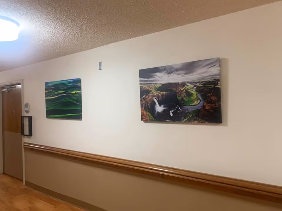 A hallway in a senior living facility with wooden handrails along the wall, two landscape paintings hanging on the wall, a closed door at the end of the hallway, and a ceiling light fixture.