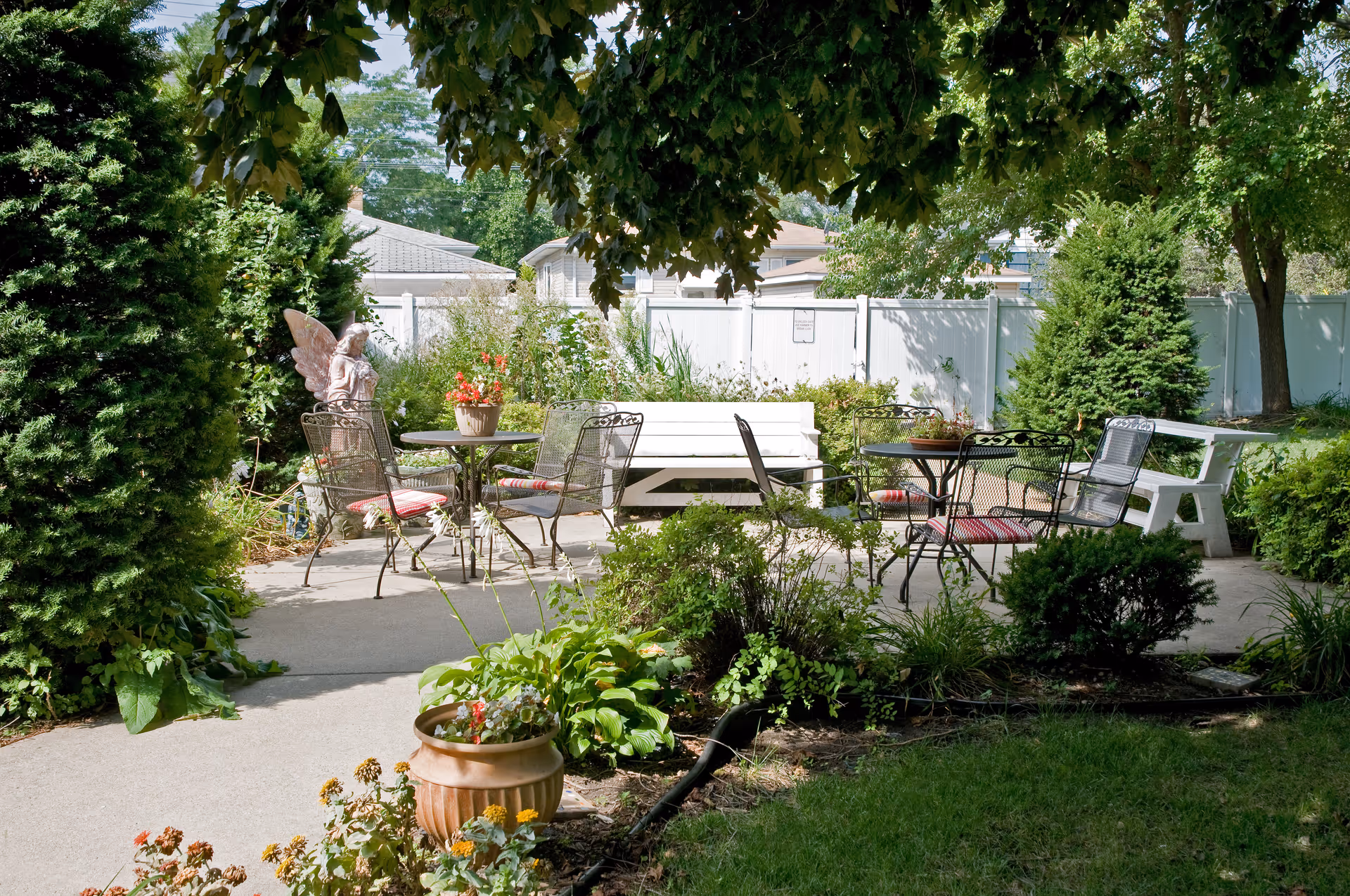Shaded outdoor patio garden with metal tables and chairs, benches, potted plants, and a decorative statue.
