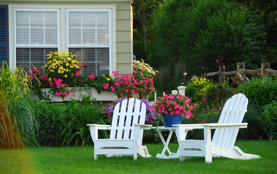 Two white Adirondack chairs and a small table on a lawn in front of a flower-filled garden and a house window.