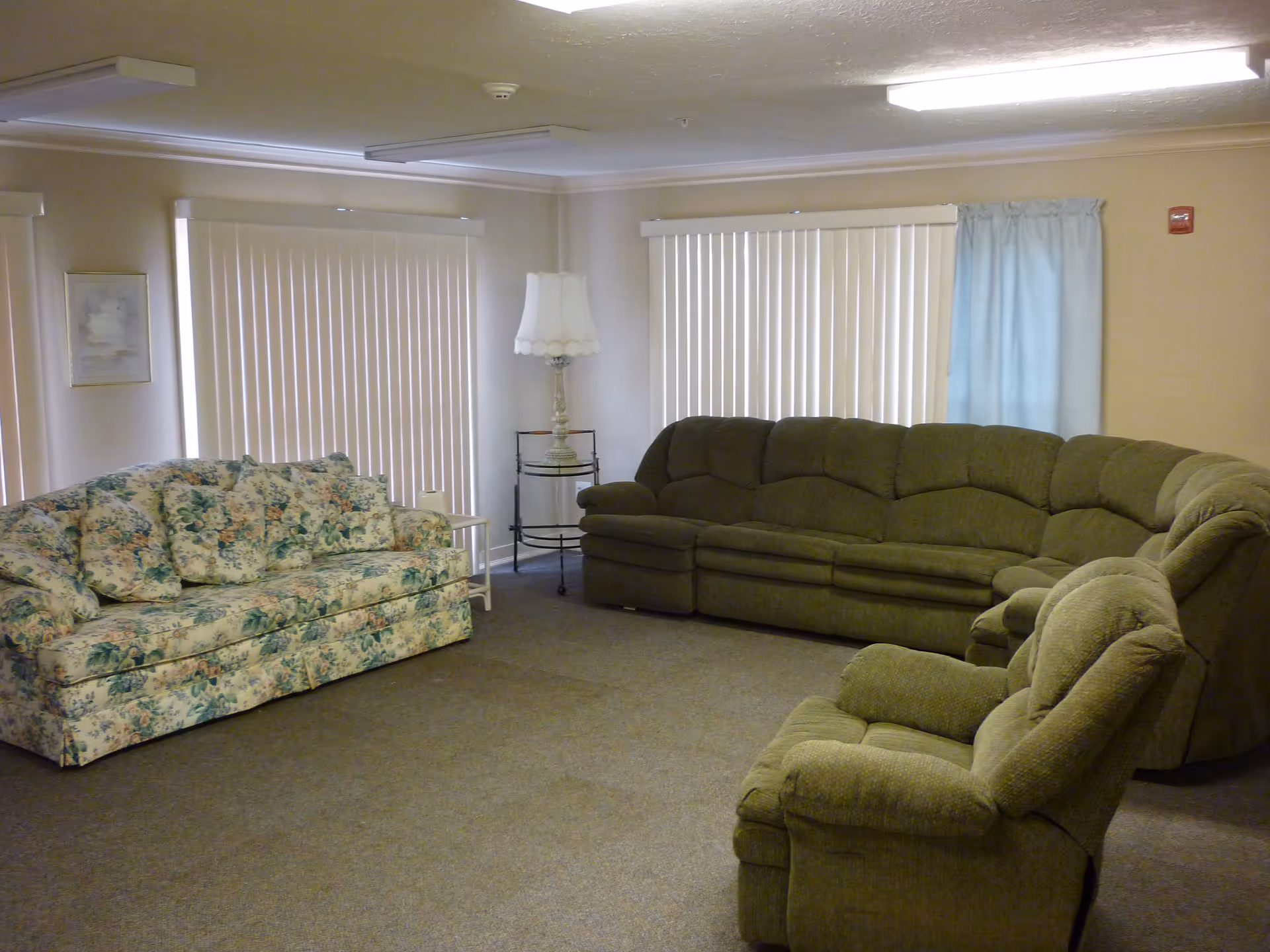 A living room with a floral patterned sofa, a green sectional sofa, and a matching green armchair. The room has beige walls, carpeted floor, vertical blinds on the windows, a standing lamp, and a small side table.