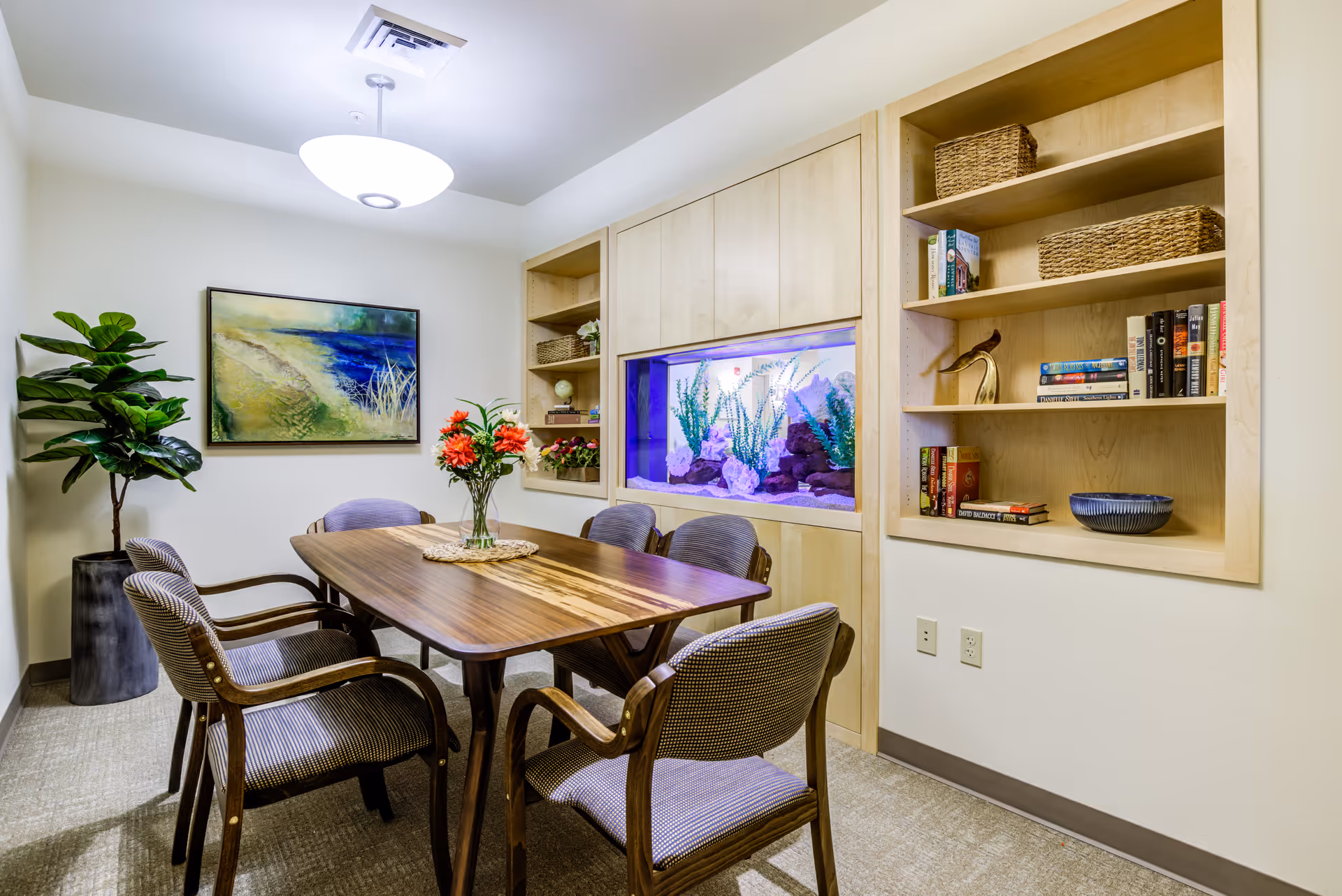 A cozy meeting or dining room with a wooden table surrounded by six cushioned chairs. The room features built-in wooden shelves with books, baskets, and decorative items. A large aquarium with plants and rocks is built into the wall. There is a potted plant in the corner and a framed painting of a beach scene on the wall. The room is well-lit with a ceiling light fixture.