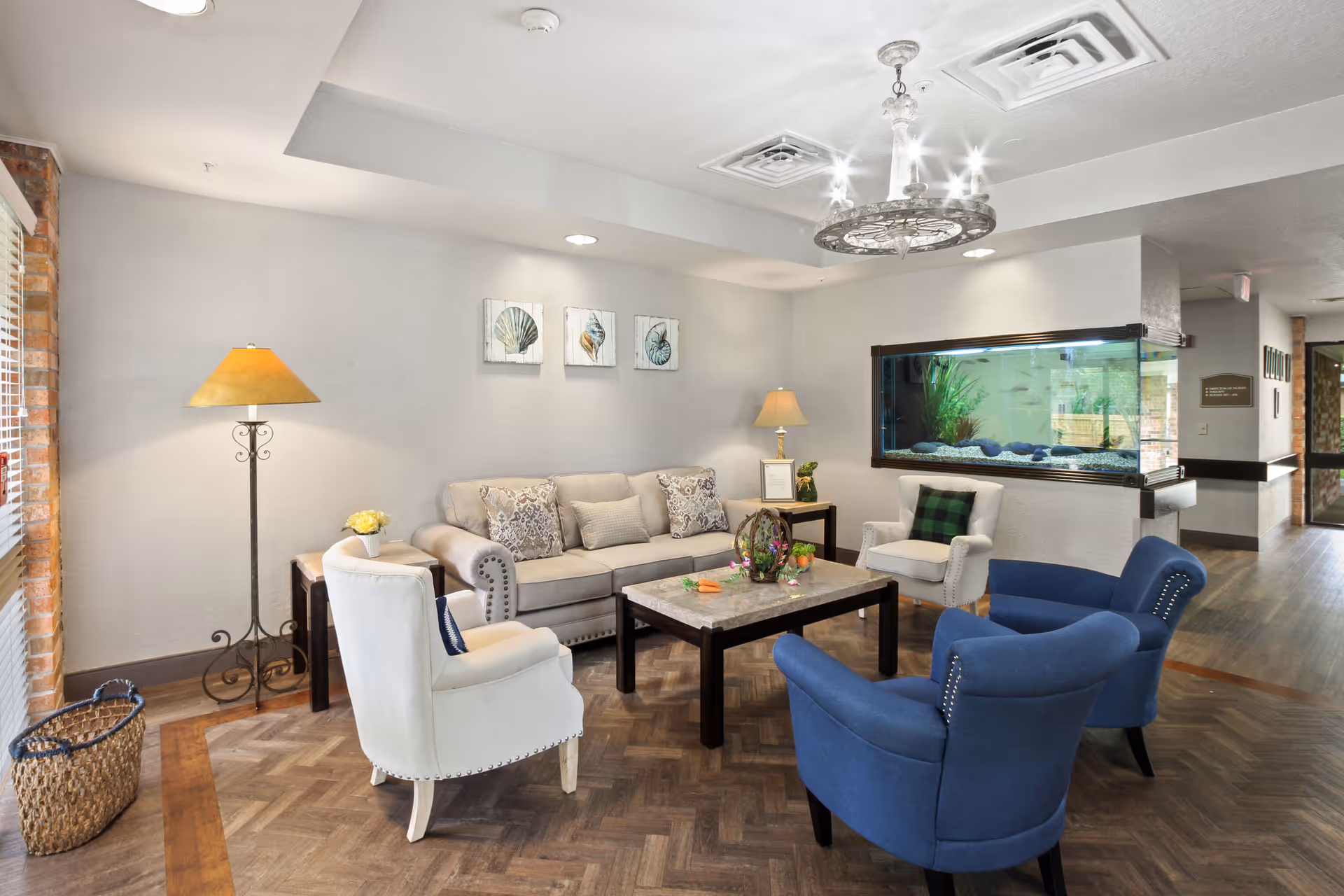 A cozy living room area in a senior living facility featuring a beige sofa with decorative pillows, two white armchairs, and two blue armchairs arranged around a wooden coffee table. The room has a floor lamp, a table lamp, three framed seashell artworks on the wall, a large built-in aquarium, and wood-patterned flooring with a basket near the window.