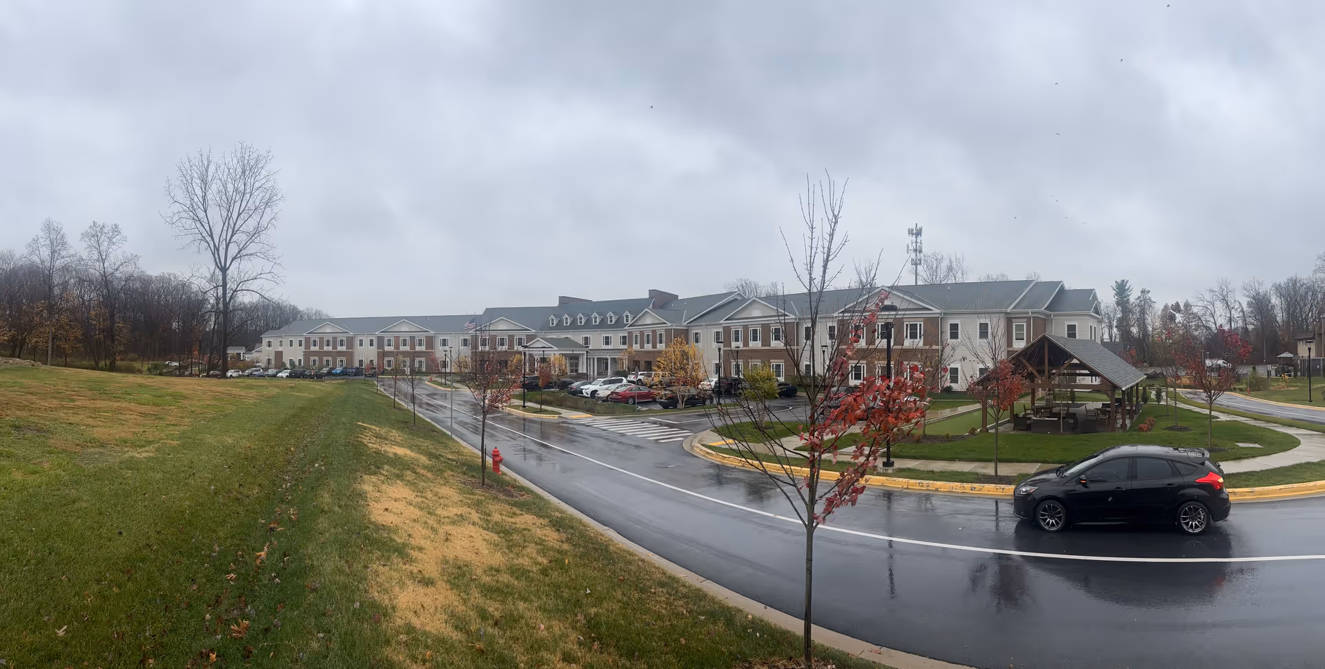 Exterior view of HarborChase of Olney senior living facility on a cloudy day with wet pavement, a black car driving on the road, a grassy area with young trees, and a covered outdoor seating area.