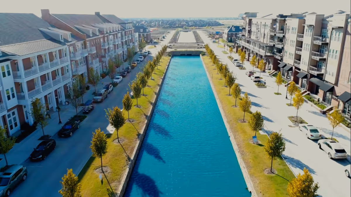 Aerial view of a long, narrow canal flanked by rows of young trees and parked cars on both sides, with multi-story residential buildings lining the streets beside the canal under a clear sky.