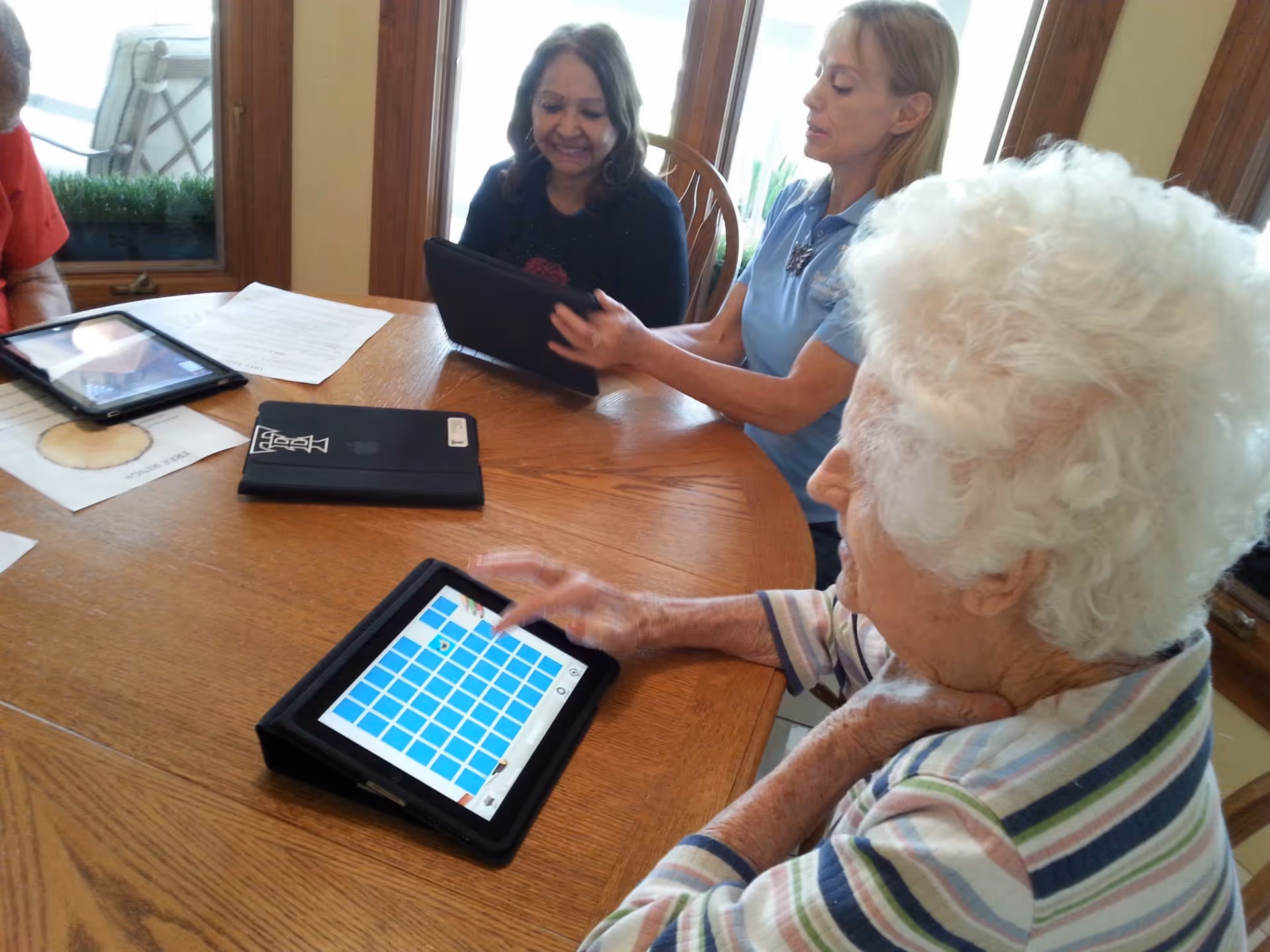 Three women sitting around a wooden table in a well-lit room, each using a tablet device. One elderly woman with white curly hair is in the foreground, interacting with a tablet displaying a grid of blue squares. Two other women, one with dark hair and one with blonde hair, are seated across the table, also holding tablets and smiling.