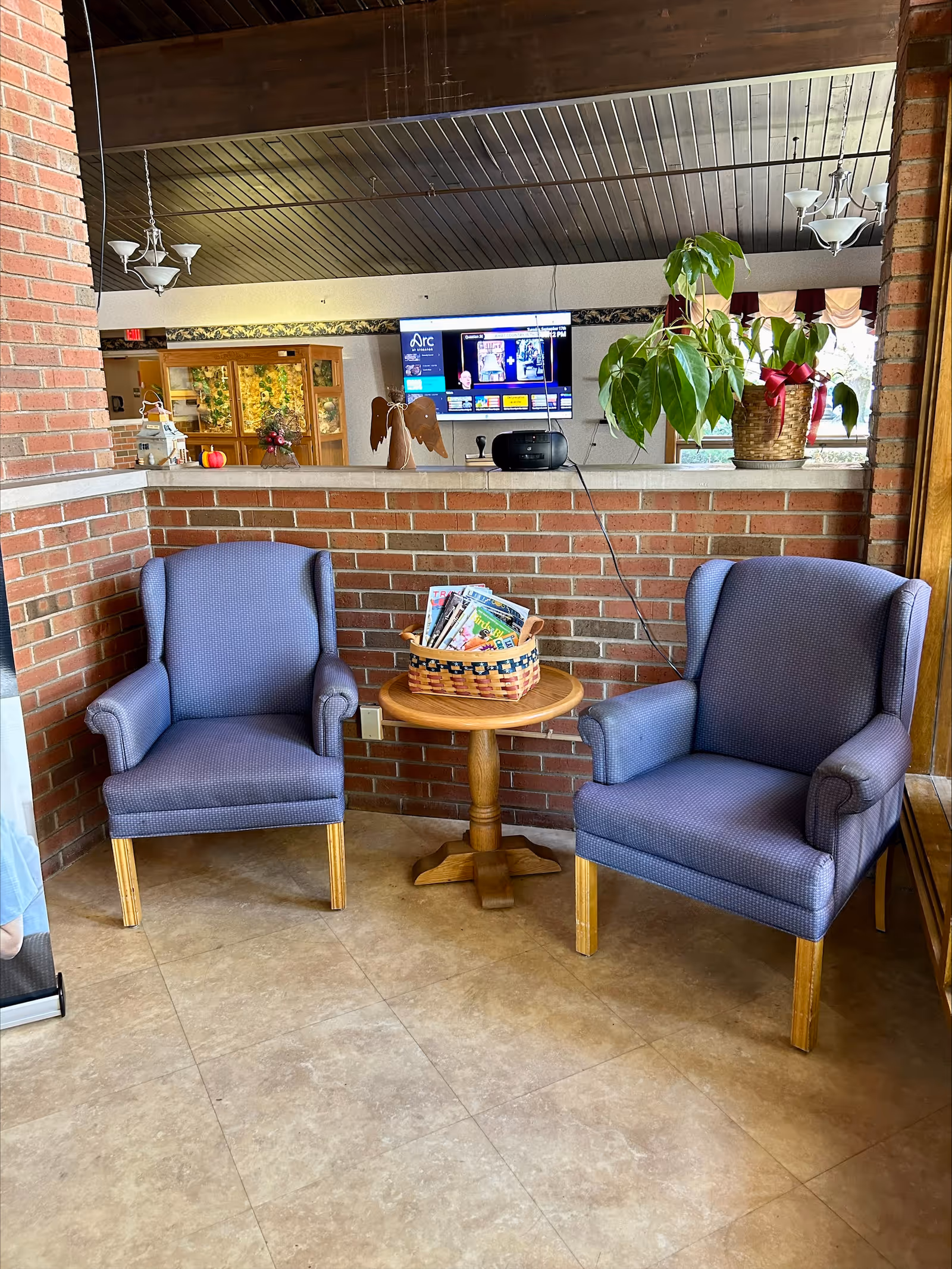 Two blue upholstered armchairs with wooden legs are positioned on either side of a small round wooden table holding a basket filled with magazines. Behind the chairs is a half-height brick wall with a potted plant and decorative items on top. A flat-screen TV is mounted on the wall in the background, and the ceiling features wooden paneling with hanging light fixtures.