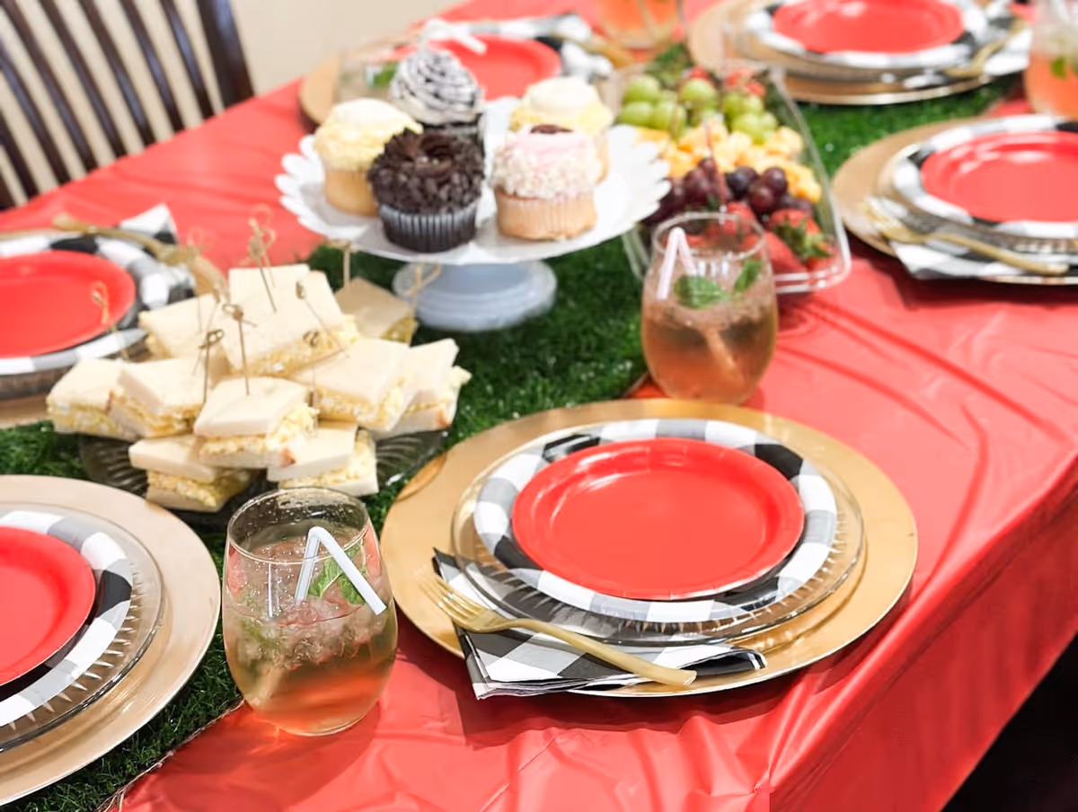 A dining table set with red plates on top of black and white checkered plates, gold chargers, and gold forks on black and white checkered napkins. The table is covered with a red tablecloth and decorated with a green grass-like runner. There are glasses with iced beverages and a variety of food including finger sandwiches, cupcakes, and a fruit platter with grapes and strawberries.